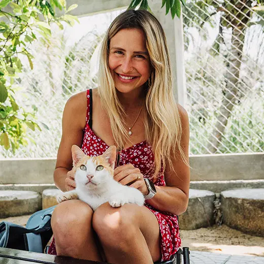 A happy smiling female visitor at Jack's Cat Cafe Hoi An, wearing a red floral dress, holds and strokes Pia, a male white and orange rescue cat, on her lap. Pia looks calmly at the camera. The background shows a sunny outdoor patio with lush green plants.