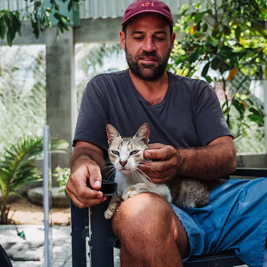 A male visitor at Jack's Cat Cafe in Hoi An gently petting Thumper, a tabby and white rescue cat, on his lap. Thumper has a relaxed expression as the guest scratches his chin. The setting is the cafe's bright, tropical outdoor area, showcasing the peaceful bond between visitors and rescue animals in Vietnam.