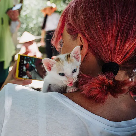 A heartwarming close-up of a visitor with vibrant red hair taking a selfie with a tiny white and ginger rescue kitten at Jack's Cat Cafe in Hoi An. The kitten is perched on the guest's shoulder, looking curiously toward the camera. In the sunlit background, other visitors wearing traditional Vietnamese conical hats (Nón Lá) can be seen, highlighting the cafe's unique local atmosphere.